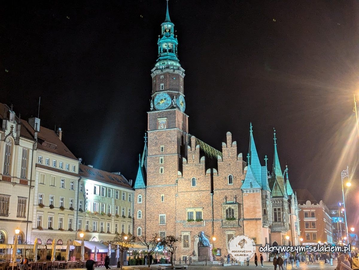 Gotycki Ratusz we Wrocławiu z wieżą i szczytami podświetlony nocą, przed nim mokra płyta Rynku
