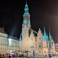 Gotisches Rathaus in Breslau mit Turm und Giebeln, nachts beleuchtet, davor das nasse Pflaster des Marktplatzes