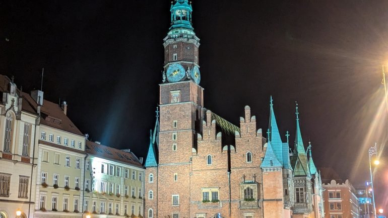 Gotycki Ratusz we Wrocławiu z wieżą i szczytami podświetlony nocą, przed nim mokra płyta Rynku
