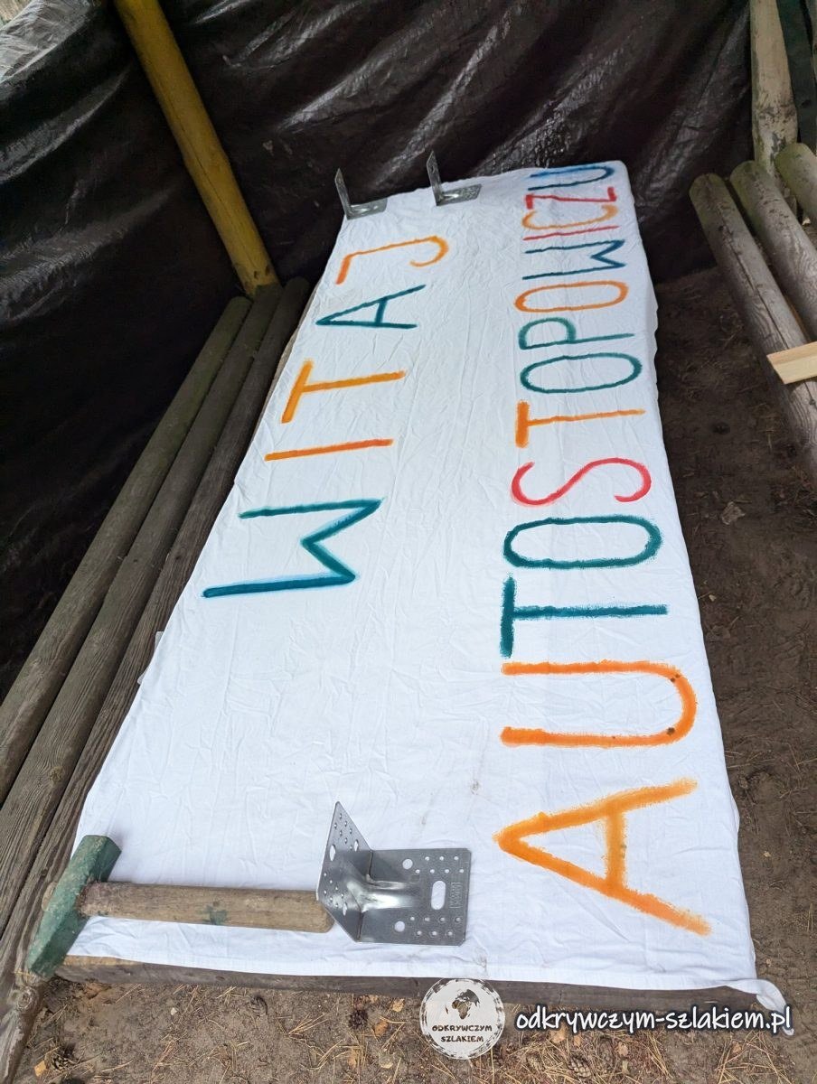 Das bemalte Banner „Witaj autostopowiczu“ (Willkommen, Tramper) auf dem Boden unter einem Unterstand