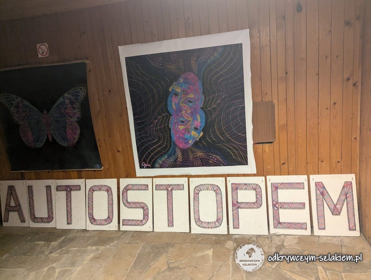 Der Schriftzug „Autostopem“ (per Anhalter) aus Buchstaben auf Pappe und zwei bunte Bilder an einer Holzwand