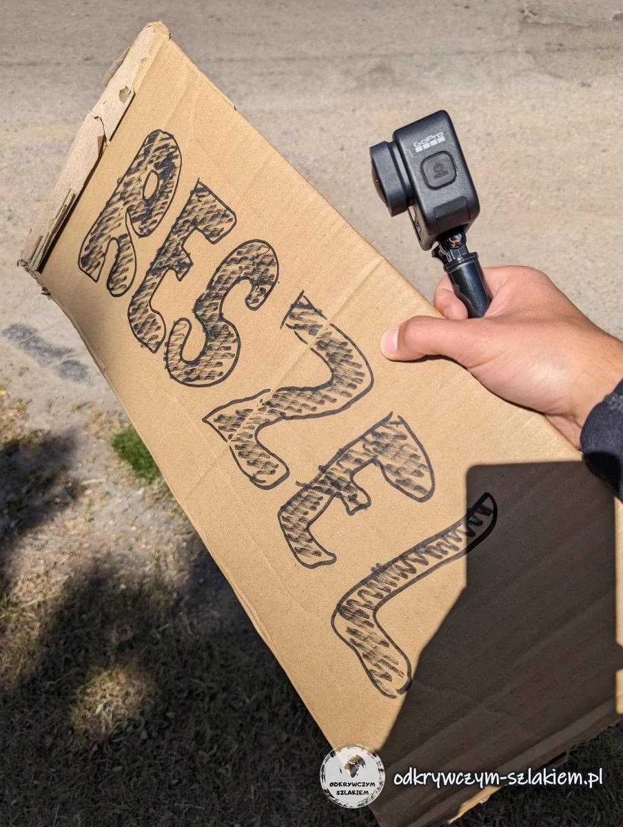 Ein Pappschild mit der Aufschrift „Reszel“ und eine GoPro-Kamera in der Hand an der Straße