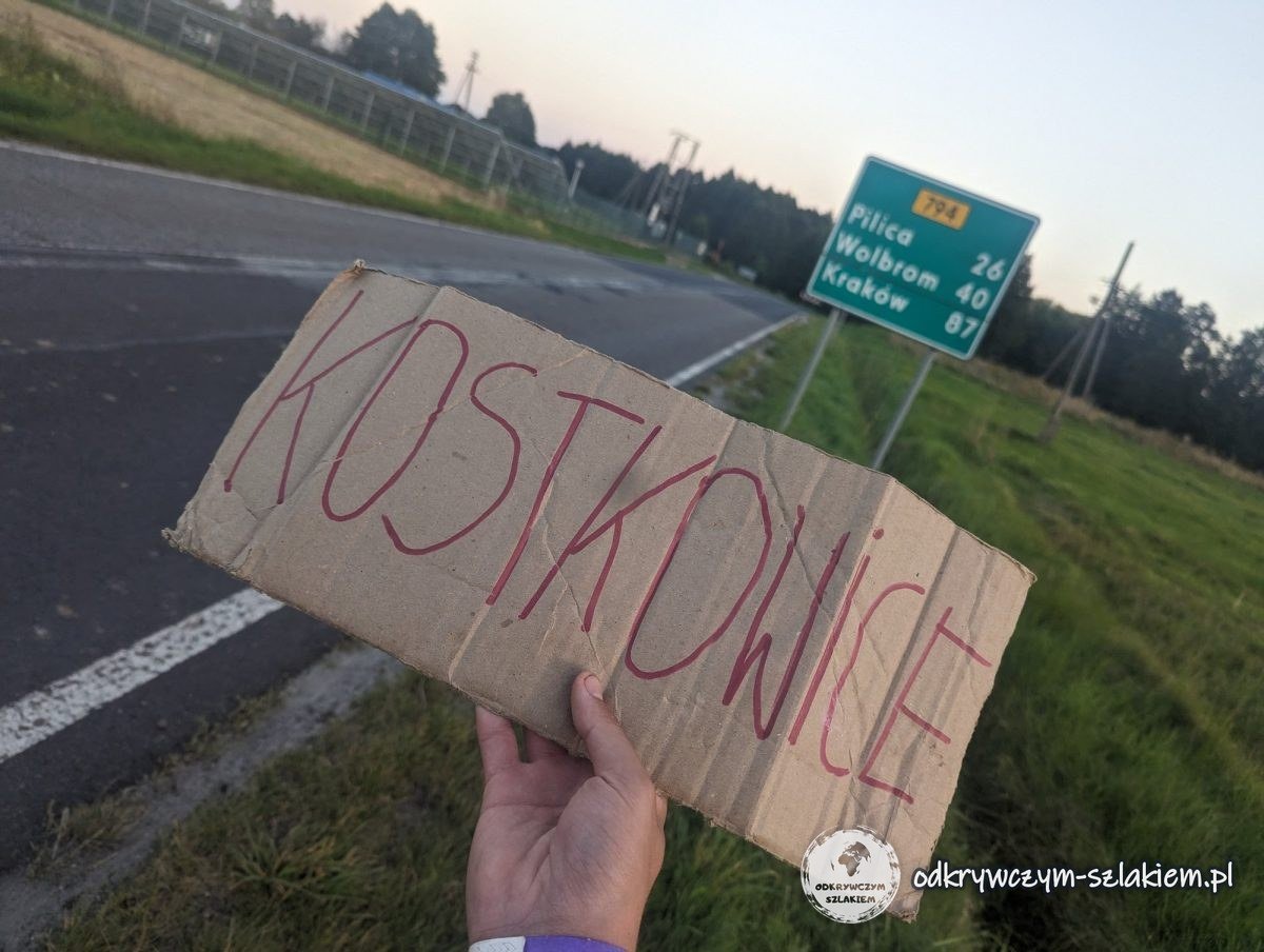Ein Schild „Kostkowice“ in der Hand an der Straße, dahinter ein Wegweiser nach Pilica, Wolbrom und Krakau
